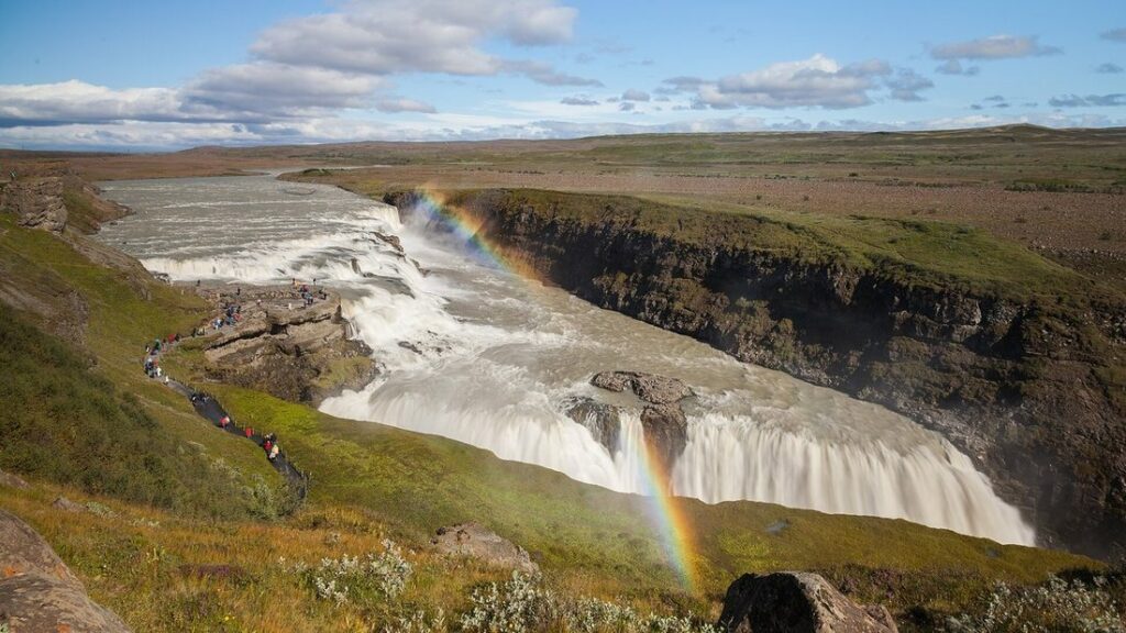 Large chute d'eau dans un canyon aux falaises sombres, avec un arc-en-ciel et un sentier de visiteurs