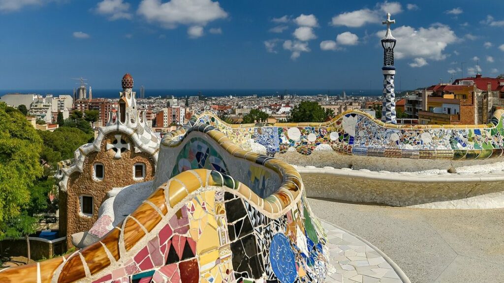Banc ondulé recouvert de mosaïques colorées au Park Güell, avec la ville de Barcelone et la mer à l'horizon