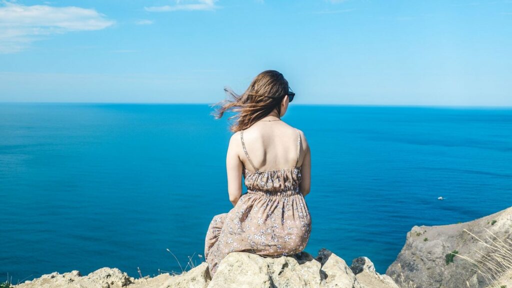 Femme assise de dos sur un rocher au bord d'une falaise, face à la mer bleue