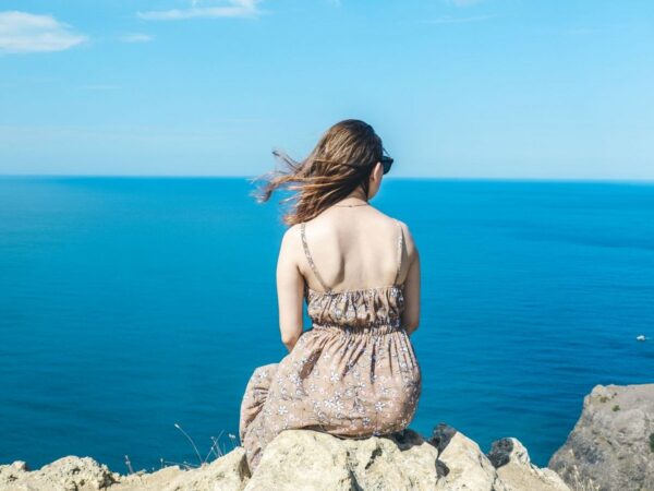 Femme assise de dos sur un rocher au bord d'une falaise, face à la mer bleue