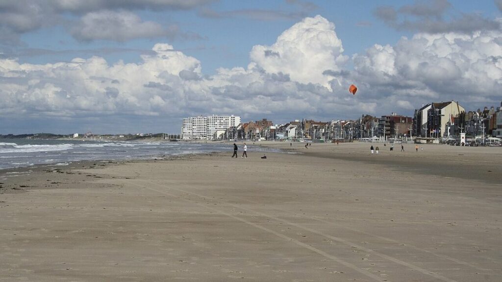 Large plage de sable à Dunkerque, vagues à gauche, front de mer au loin et cerf-volant orange dans le ciel