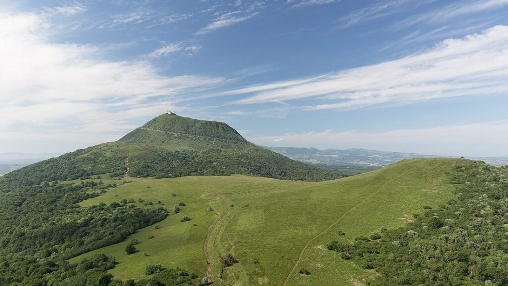 Le puy de Dôme, cône volcanique herbeux surmonté d'une antenne, vu d'une crête verte sous un ciel strié de nuages