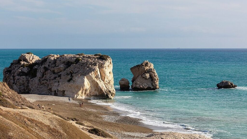 Gros rocher clair et deux plus petits dans une mer turquoise, devant une plage de galets
