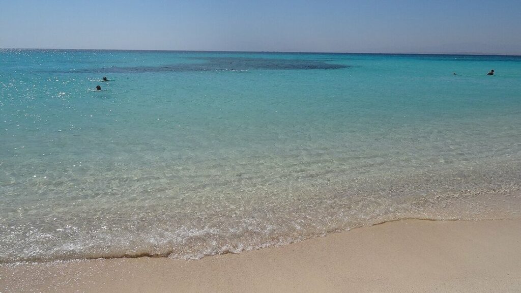 Eau turquoise très claire et transparente qui lèche une plage de sable fin, quelques baigneurs au loin