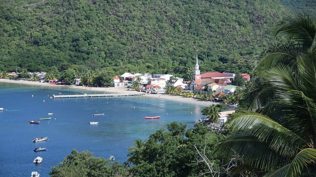 Baie bordée d'une plage et d'un village dominé par un clocher, sous une colline boisée, petits bateaux sur l'eau bleue