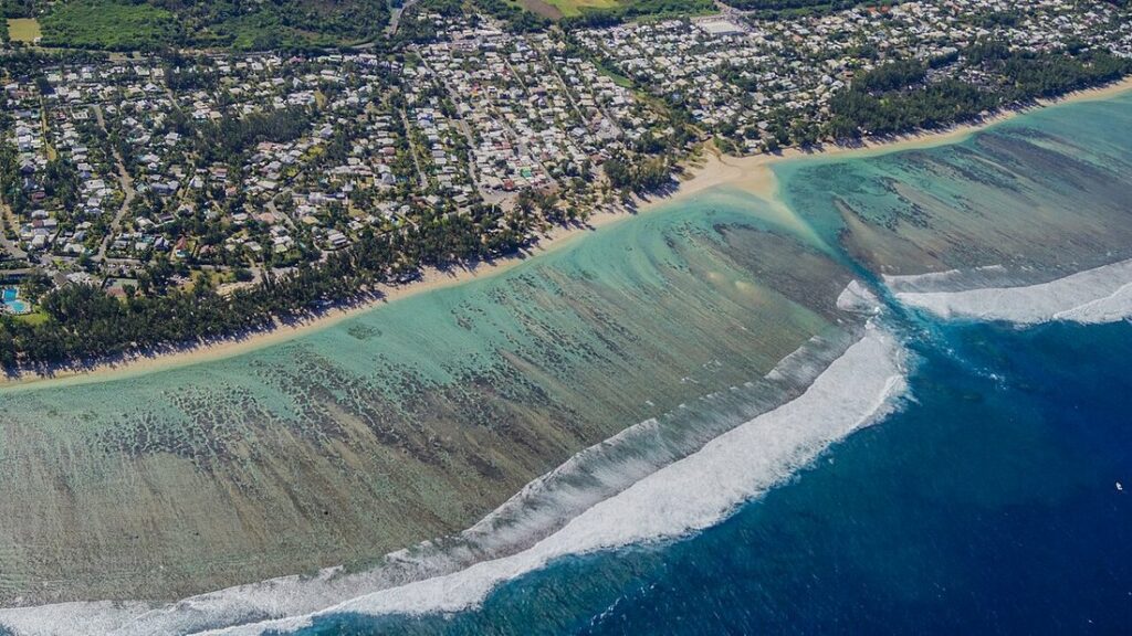 Vue aérienne d'un récif corallien turquoise frangé de vagues blanches, devant une plage et un quartier résidentiel arboré