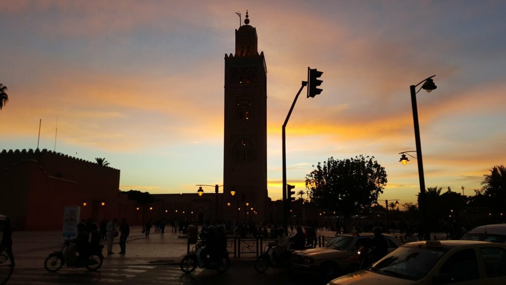 Silhouette du minaret de la Koutoubia à Marrakech sur un ciel orangé au crépuscule, scooters et taxis au premier plan