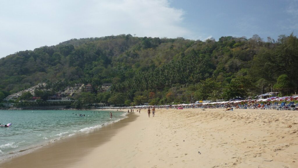 Plage de sable clair bordée de collines boisées, eau turquoise, baigneurs et rangées de parasols, plage de Nai Harn à Phuket