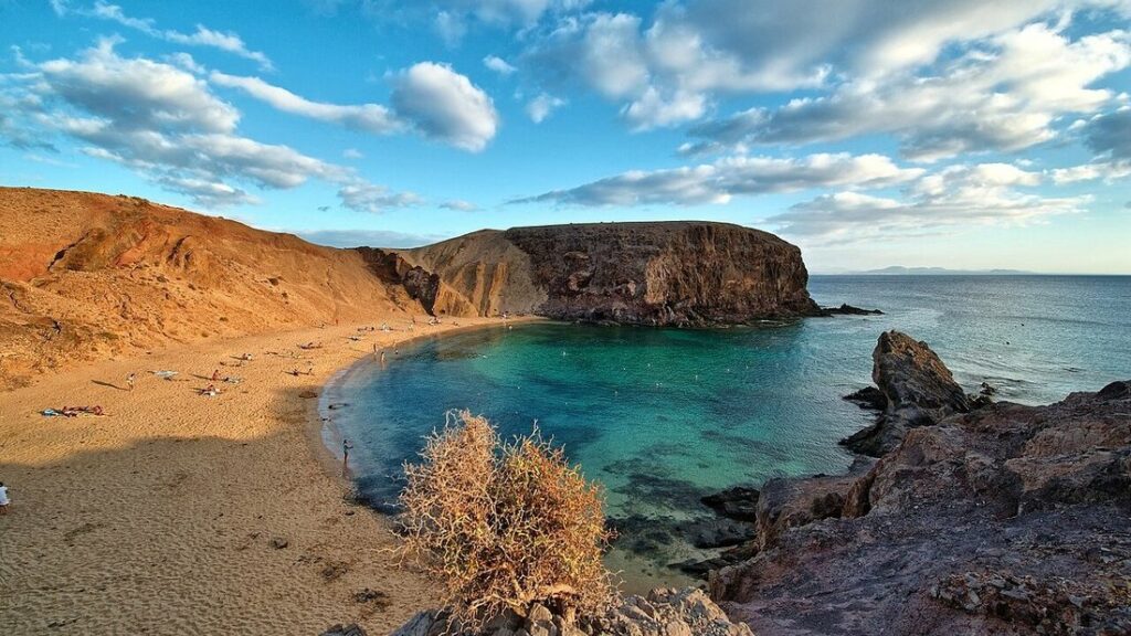Crique de sable ocre entourée de falaises rousses, eau turquoise et ciel nuageux, plage de Papagayo à Lanzarote