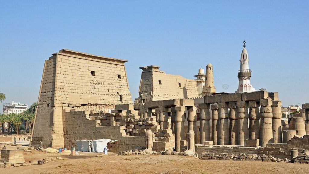 Pylône et colonnade en pierre ocre du temple de Louxor sous un ciel bleu, avec un minaret blanc à droite