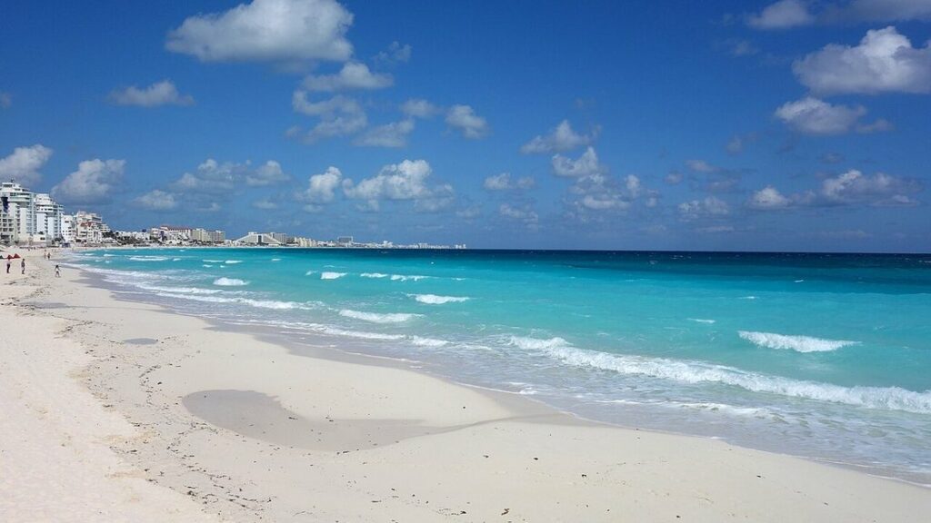 Longue plage de sable blanc et eau turquoise sous un ciel bleu parsemé de nuages, immeubles au loin à gauche