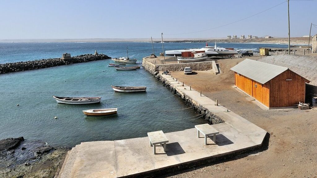 Petit port de pêche aux barques blanches sur une eau vert turquoise, jetée en béton et cabane en bois orange