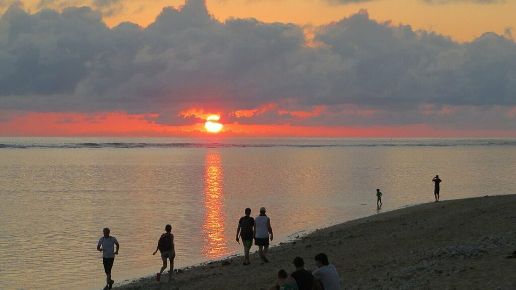 Soleil bas sous un banc de nuages sombres, reflets orangés sur une mer calme, promeneurs en silhouette sur la plage