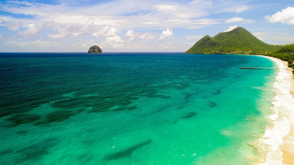 Vue aérienne d'une plage de sable clair, d'une mer turquoise, d'un îlot rocheux au large et d'une colline verte