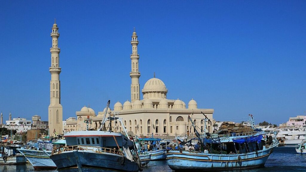 Mosquée beige à deux minarets derrière des bateaux de pêche bleus amarrés dans un port, sous un ciel bleu
