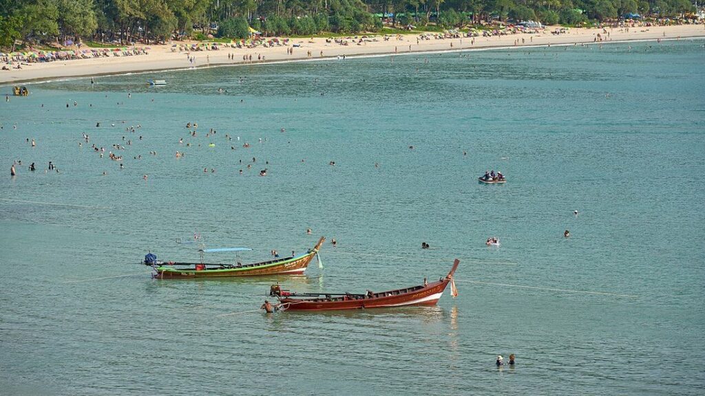 Baie de sable clair bordée d'arbres et de parasols, avec deux bateaux longue queue sur une eau turquoise