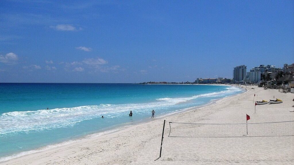 Longue plage de sable blanc face à une mer turquoise à vagues, avec des immeubles au loin et un filet de volley