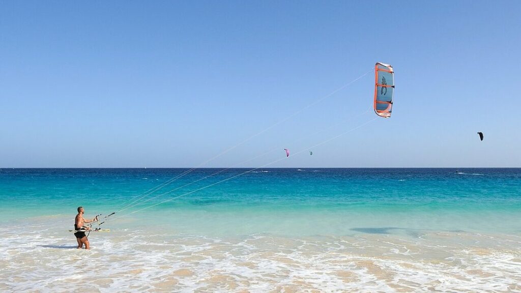 Kitesurfeur dans l'eau peu profonde, tenant les lignes d'une grande aile orange et bleue, devant une mer turquoise
