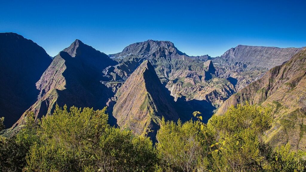 Crêtes et pitons montagneux aux pentes ocre et vertes sous un ciel bleu, avec de la végétation au premier plan