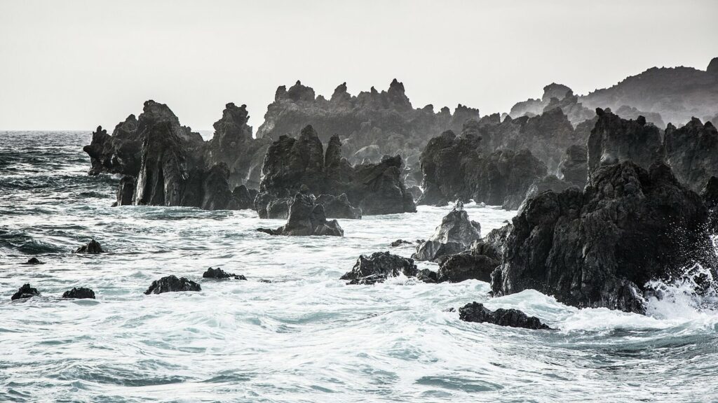 Rochers de lave noire battus par de grosses vagues écumantes sous un ciel gris pâle, sur une côte de Lanzarote