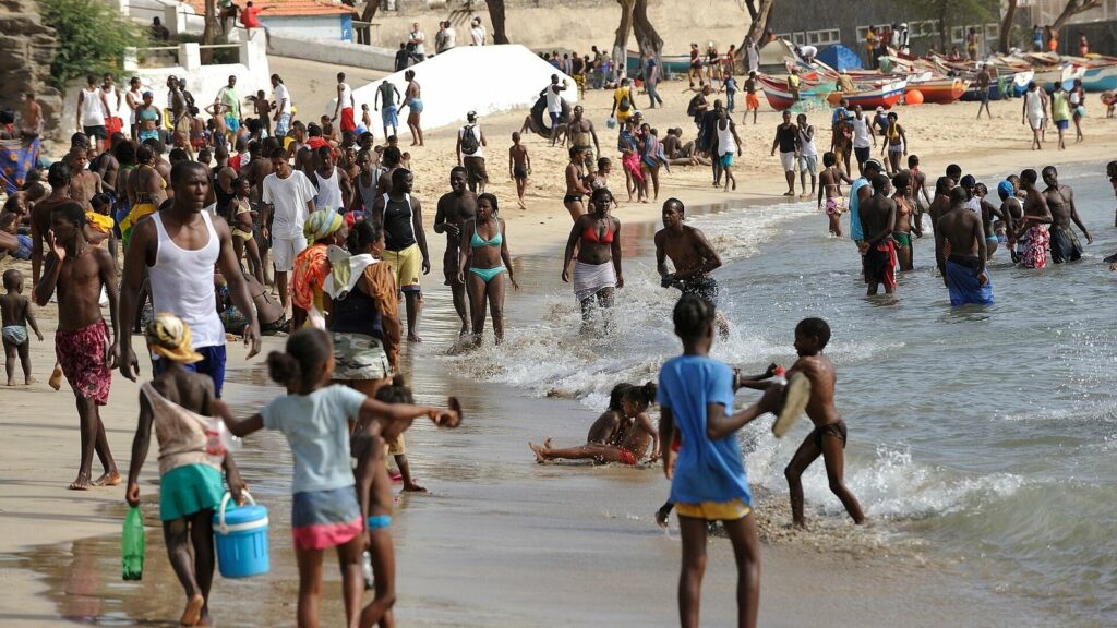 Plage de sable animée à Praia au Cap-Vert, avec des baigneurs dans l'eau, des enfants au bord des vagues et des barques de pêche colorées tirées sur le sable