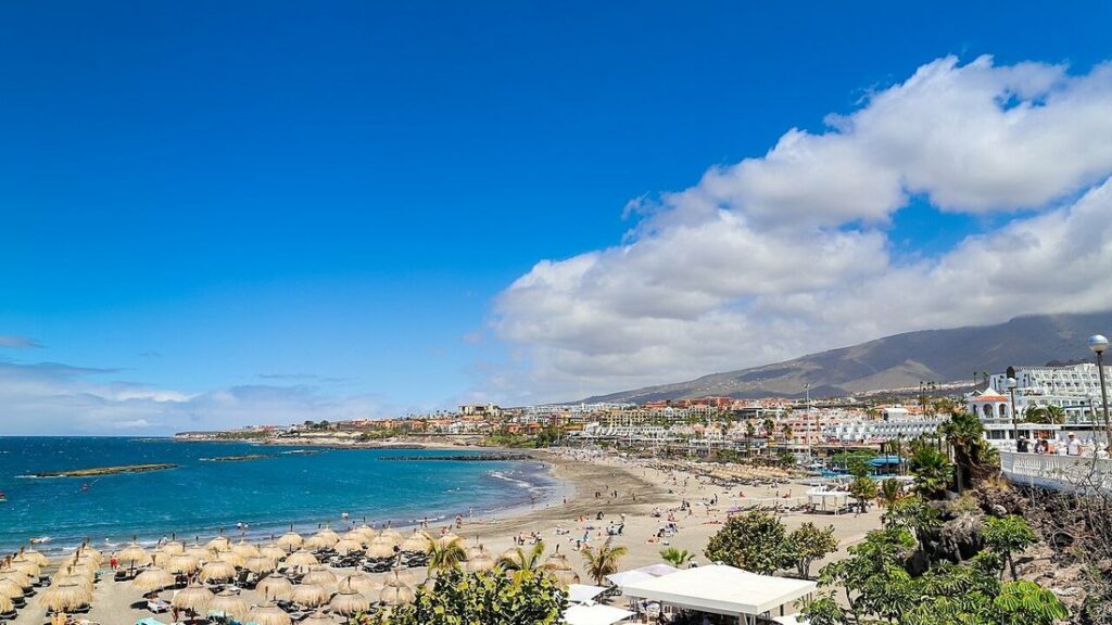 Plage de sable avec parasols de paille, eau turquoise et front de mer bâti au pied d'une montagne brune