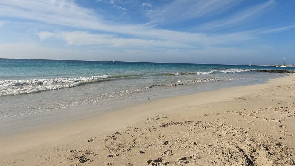 Plage de sable clair avec petites vagues, mer turquoise et voiliers à l'horizon