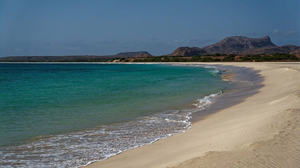 Long croissant de sable clair au bord d'une eau turquoise, avec des collines brunes au loin