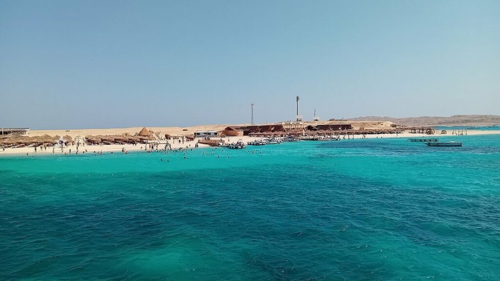 Eau turquoise devant une île de sable plate avec paillotes, bateaux et baigneurs au loin