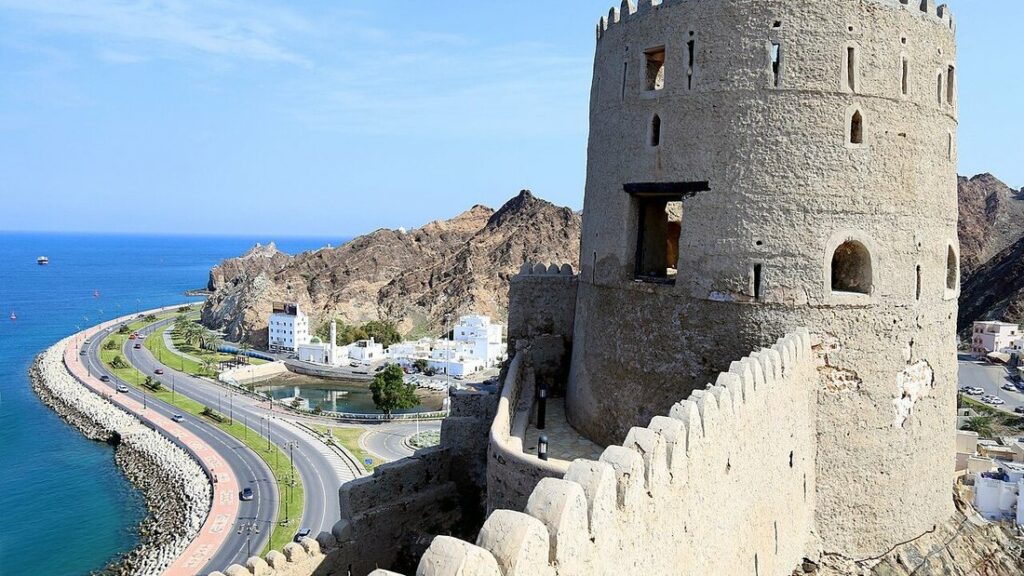Tour crénelée d'un fort dominant une route en bord de mer, des maisons blanches et des montagnes rocheuses
