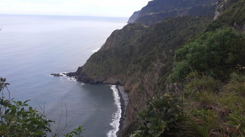 Falaises couvertes de végétation tombant dans l'océan, au-dessus d'une plage de galets sombres, sous un ciel voilé