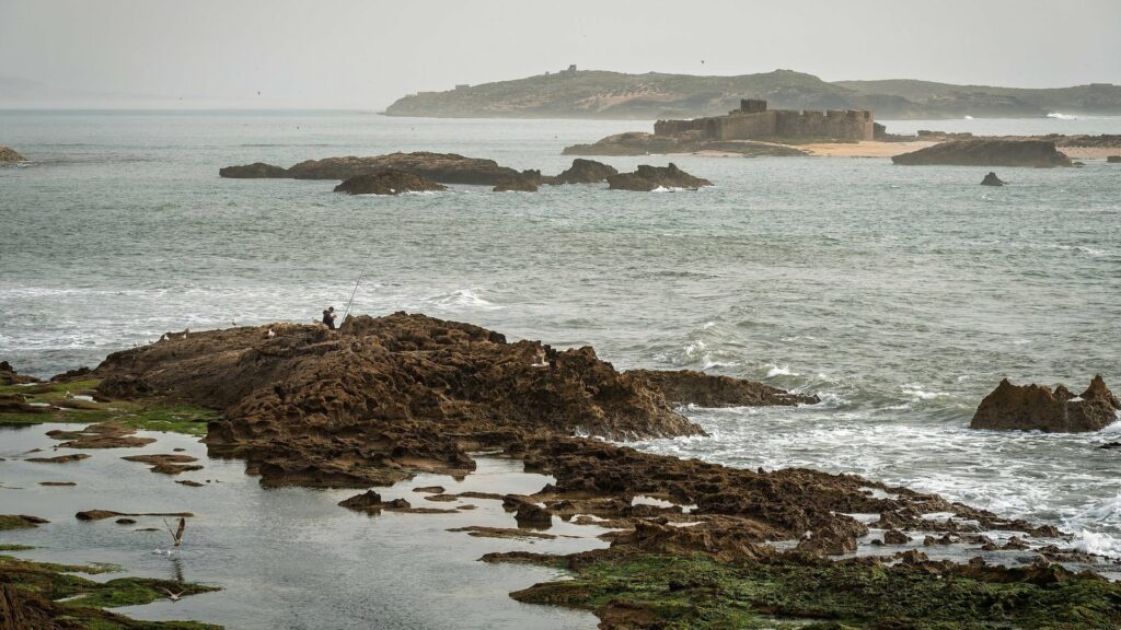 Rochers bruns creusés par la mer au bord d'un océan gris, avec un pêcheur à la ligne et, au loin, un îlot portant les vestiges d'un fort, sous un ciel brumeux