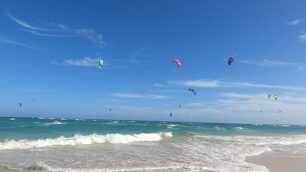 Nombreux kites de kitesurf colorés dans un ciel bleu, au-dessus d'une mer turquoise et de vagues écumeuses