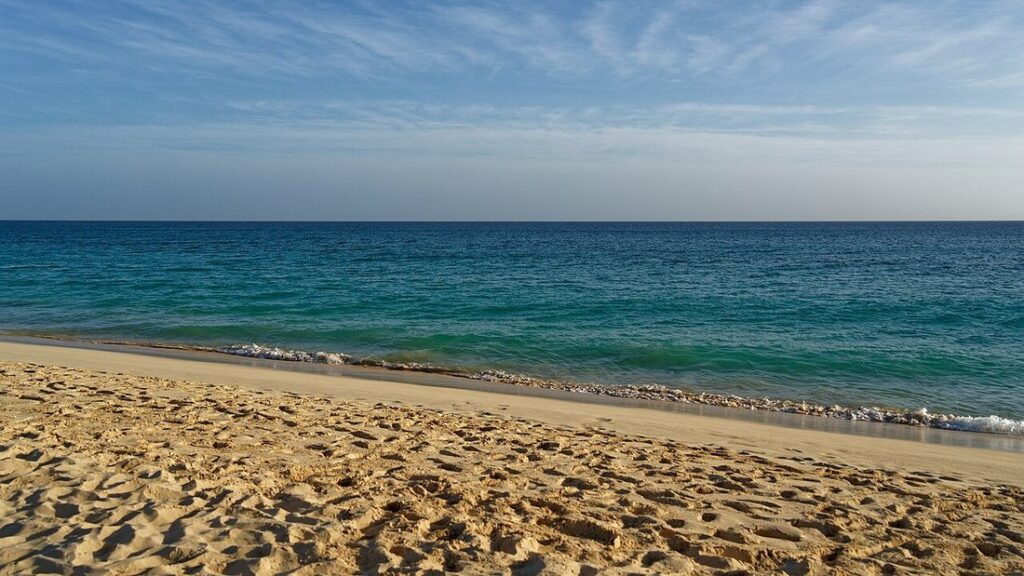 Plage de sable doré marquée de traces de pas, devant une mer bleu-vert et un ciel voilé