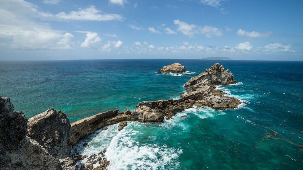 Pointe rocheuse s'avançant dans une mer turquoise, avec des îlots, de l'écume et une île au loin