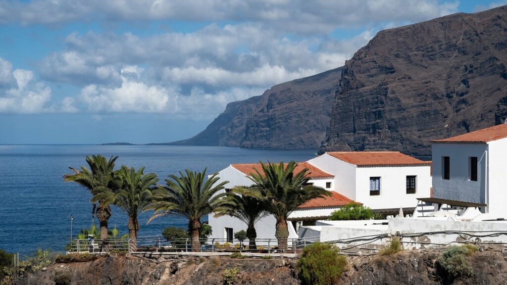 Palmiers et maisons blanches aux toits de tuiles rouges devant l'océan et les hautes falaises de Los Gigantes, à Tenerife