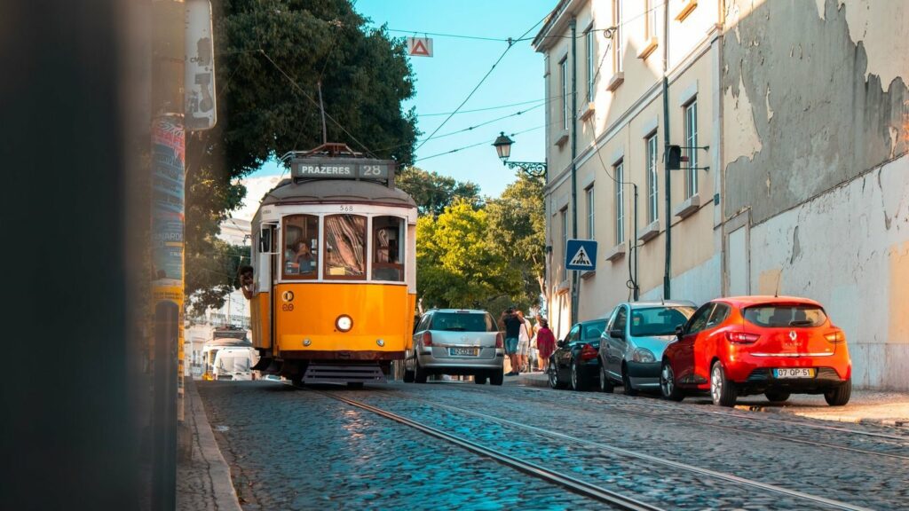Tramway jaune de la ligne 28 en direction de Prazeres sur une rue pavée, avec des voitures garées le long d'un immeuble défraîchi