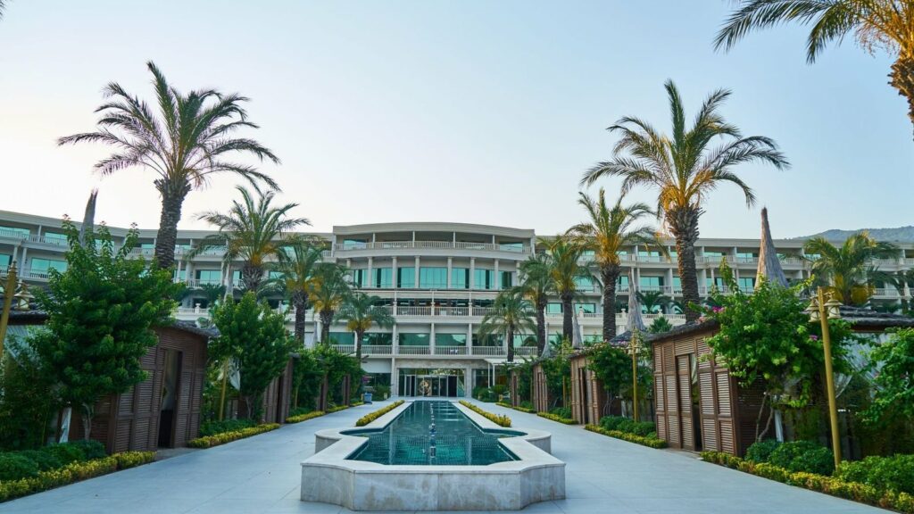Façade d'un grand hôtel de plusieurs étages entourée de palmiers, avec un bassin allongé à jets d'eau et des cabanons en bois au premier plan