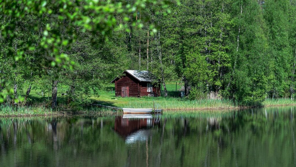 Petite cabane en rondins rouge au bord d'un lac entouré de bouleaux, avec une barque blanche amarrée