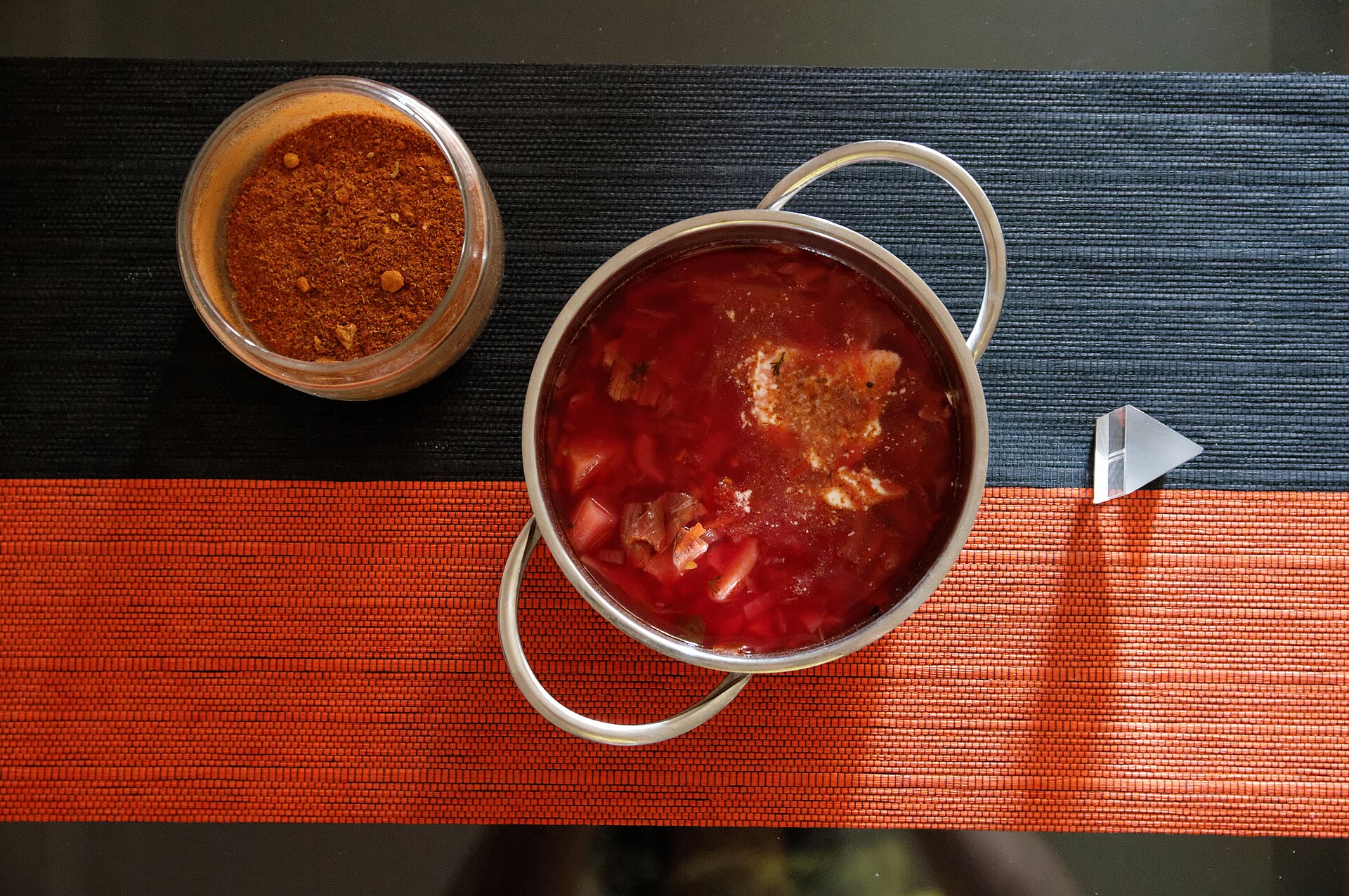 Casserole en inox remplie de soupe rouge de type bortsch, avec un bocal de poudre d'épices posé à côté, sur un set noir et orange