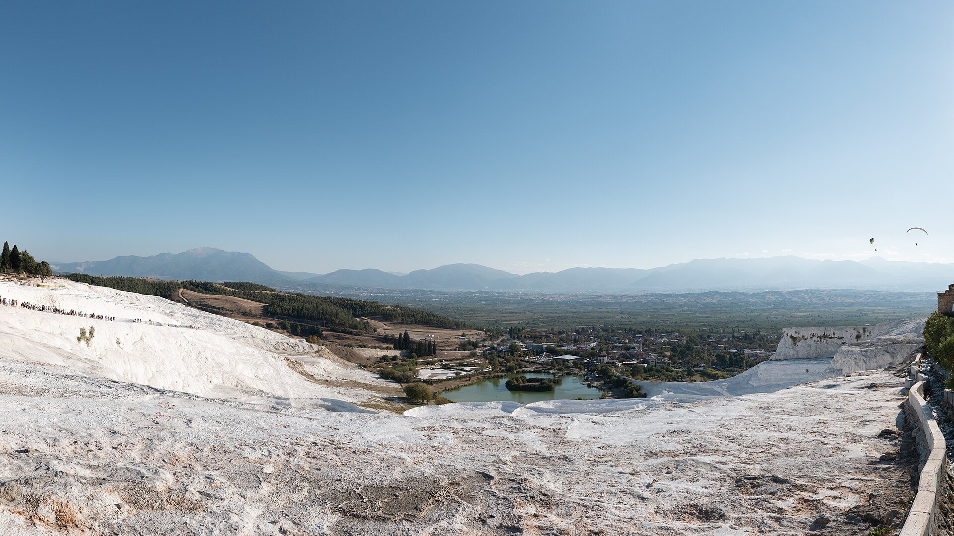 Terrasses de travertin blanc dominant une plaine, avec un village, un petit lac et deux parapentes dans le ciel
