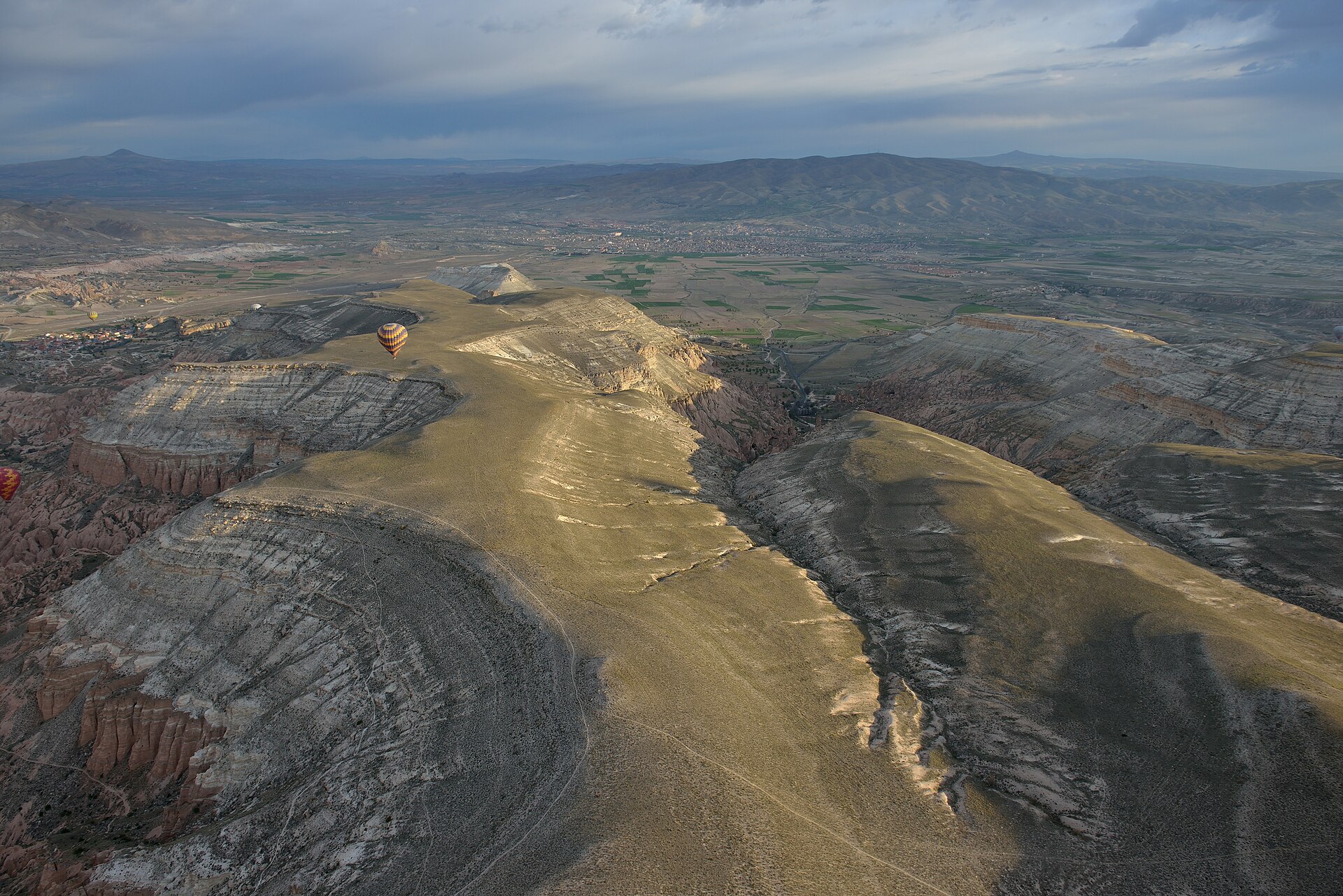 Plateaux de roche claire entaillés de canyons vus d'en haut, avec une montgolfière et une ville dans la plaine